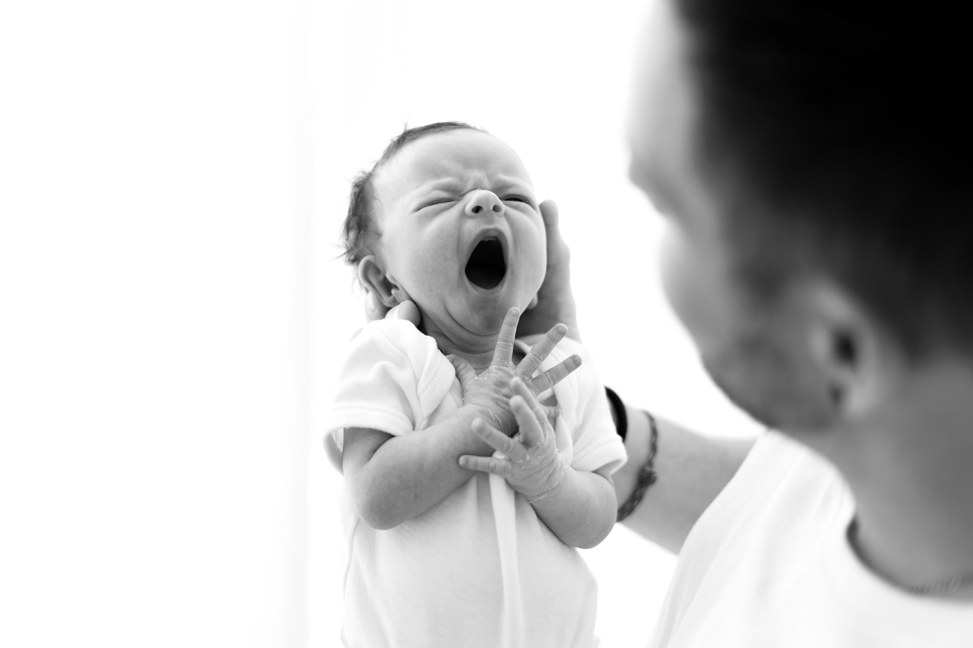 baby held in fathers hands is yawning - black and white image captured by norwich newborn photographer