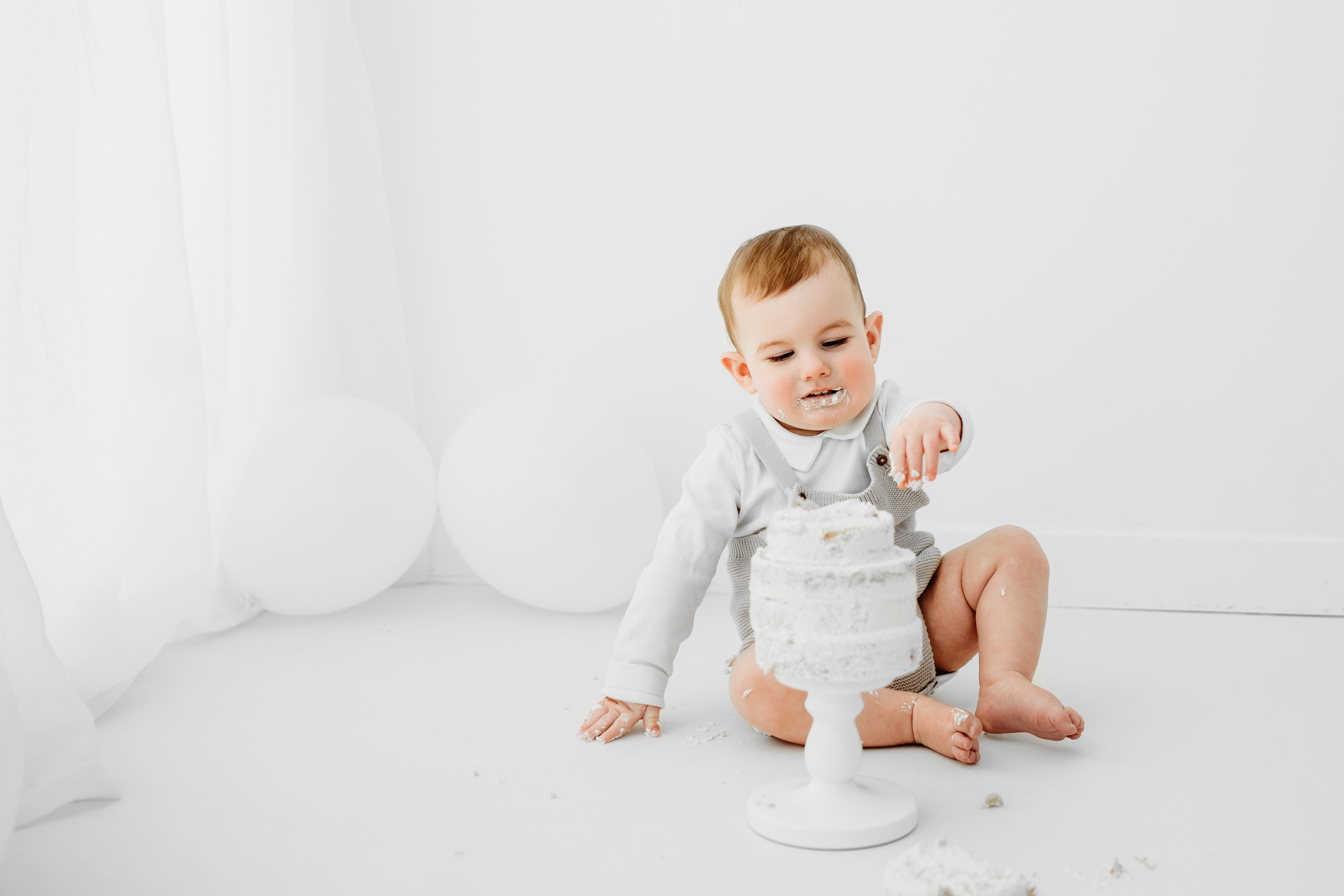 little boy sits in white studio playing with a white cake during cake smash in norwich
